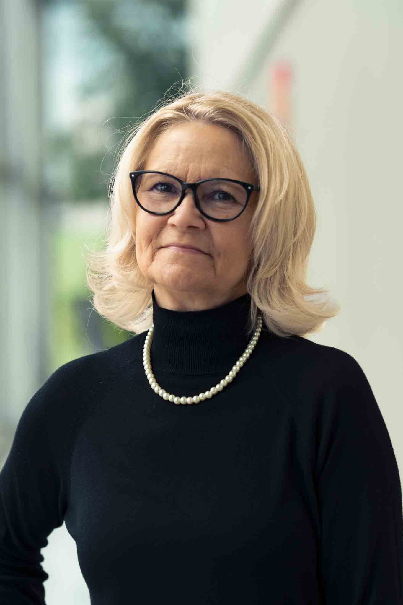 Frau in dunklem Rollkragenpullover mit hellem, schulterlangem Haar mit Brille und Perlenkette