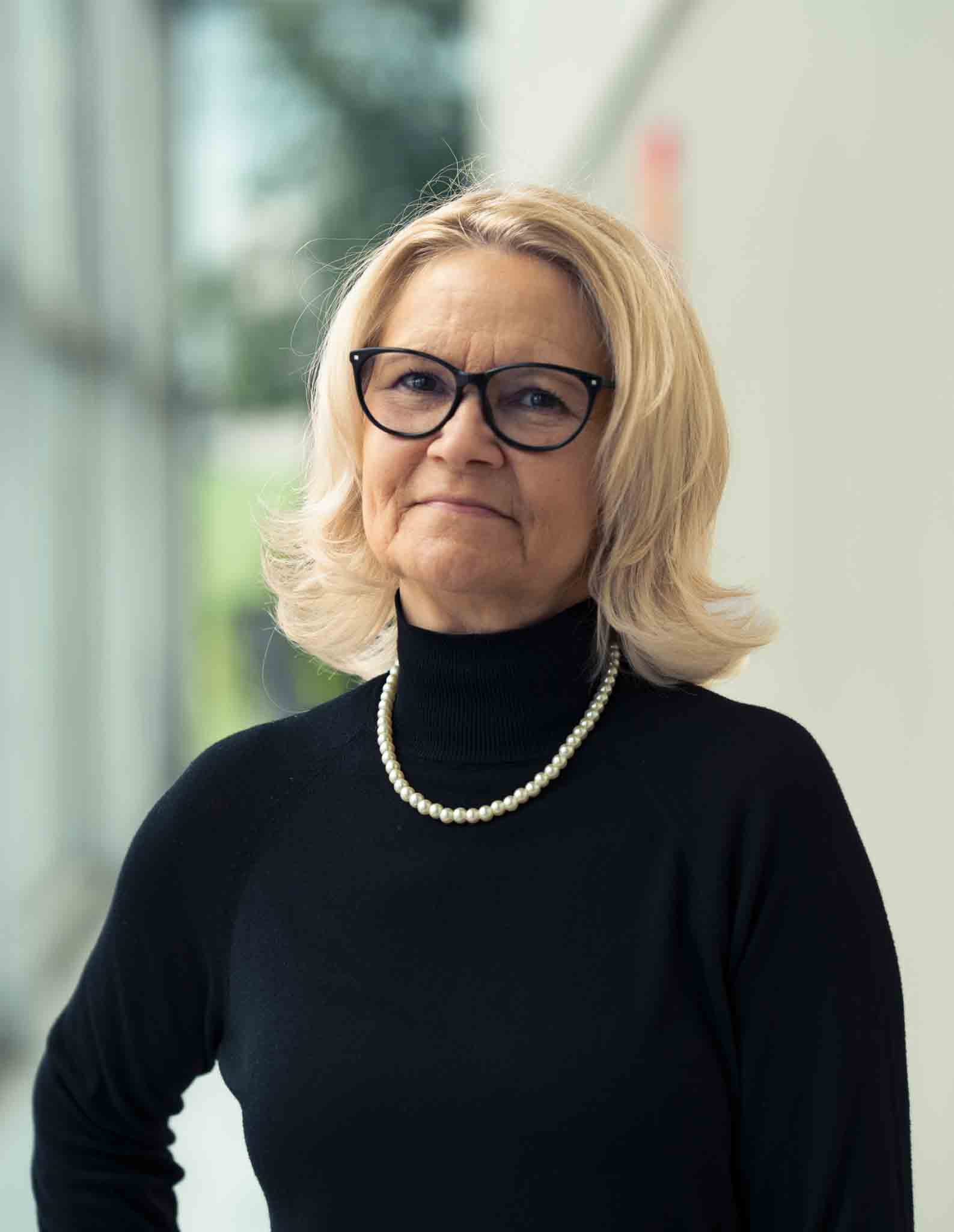 Frau in dunklem Rollkragenpullover mit hellem, schulterlangem Haar mit Brille und Perlenkette